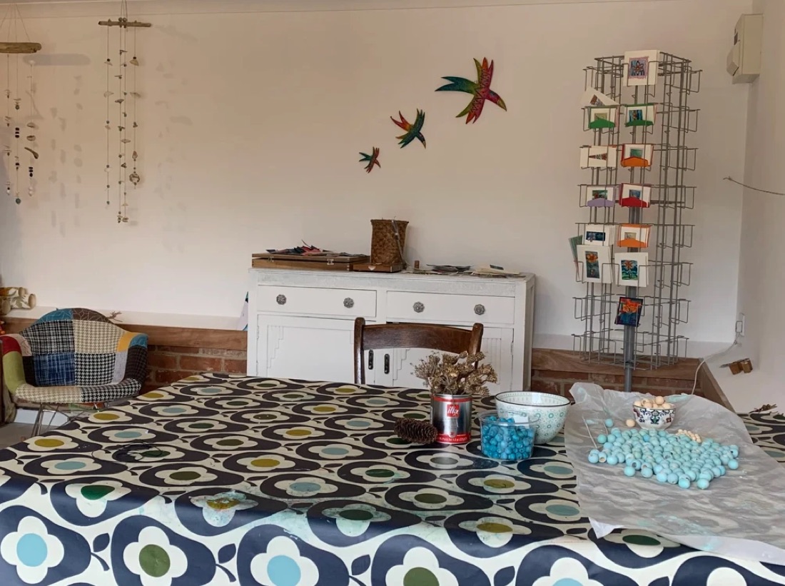The inside of the studio: a big patterned table, a patchwork chair, hanging mobiles, painted birds on the wall and a rack of handmade cards